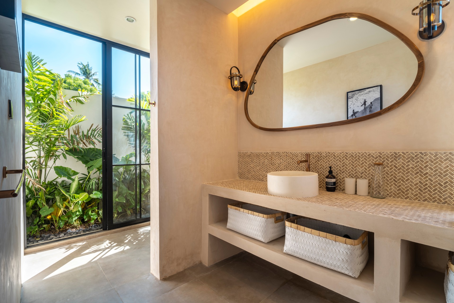 A bathroom at Santika Villas, with a curved mirror above a stone basin and a doorway open to the garden